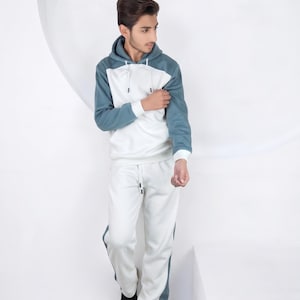 Puede incluir: Conjunto deportivo de dos piezas en blanco y verde azulado. La sudadera con capucha tiene mangas verde azulado y un cuerpo blanco. Los pantalones de chándal a juego tienen rayas verde azulado en los laterales. El conjunto se completa con zapatillas deportivas negras.