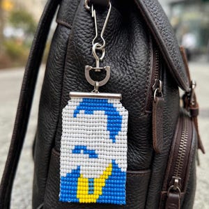 Puede incluir: Un llavero rectangular de cuentas con un diseño de retrato pixelado en blanco, azul y amarillo. El accesorio está sujeto a un cierre giratorio de metal plateado y una barra horizontal, colgado de una mochila de cuero marrón texturizado.