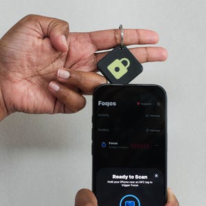 May include: A black square NFC tag featuring a light green padlock icon on a metal keyring, positioned near a smartphone displaying the Foqos app interface with a Ready to Scan prompt that instructs users to hold the phone near an NFC tag.