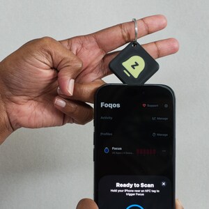 Puede incluir: Un smartphone negro que muestra la interfaz de la aplicación Foqos con un aviso de Ready to Scan. Una etiqueta NFC cuadrada negra con un icono de campana verde claro y la letra Z está unida a un llavero de metal plateado cerca de la pantalla.