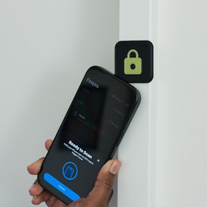 May include: A black smartphone displaying a screen that reads Ready to Scan and Hold your iPhone near an NFC tag to trigger Focus, positioned next to a square black NFC tag featuring a light green padlock icon attached to a white door frame.