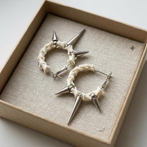 Puede incluir: Un par de pendientes de aro que presentan una serie de segmentos óseos de color blanco roto alternados con puntas de metal plateado pulido. Los pendientes tienen forma circular e incluyen un mecanismo de cierre de poste con bisagra.