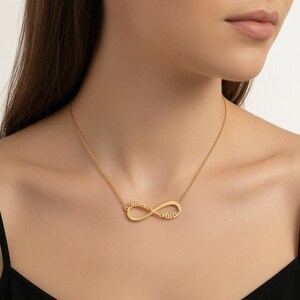 Op de afbeelding: Een goudkleurige ketting met een hanger in de vorm van een oneindigheidssymbool. De hanger is geïntegreerd in een fijne ketting en toont twee namen in cursief schrift, één op elke lus van het oneindigheidssymbool voor een persoonlijk sieraad.