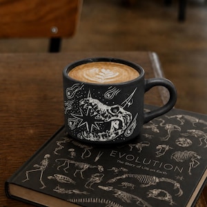 May include: A matte black ceramic mug featuring a white line art illustration of a prehistoric skull surrounded by stars and comets, filled with a latte. The mug rests on a black book titled Evolution: The Story of Life on Earth.
