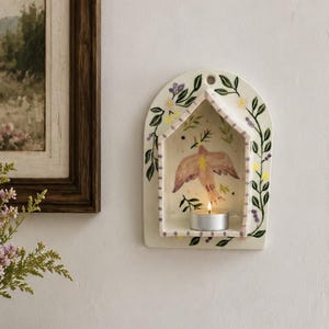 Op de afbeelding: Een crèmekleurige keramische wandkandelaar in de vorm van een huis met een boogvormige bovenkant. Het object is voorzien van handgeschilderde groene ranken, paarse bessen en gele bloemen, met een centrale illustratie van een vogel en een brandend waxinelichtje.