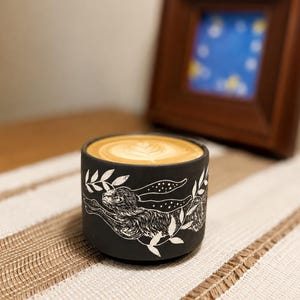 May include: A matte black ceramic mug featuring a white engraved illustration of a running hare surrounded by leafy branches. The mug is filled with a coffee beverage topped with a white foam latte art design in a circular pattern.