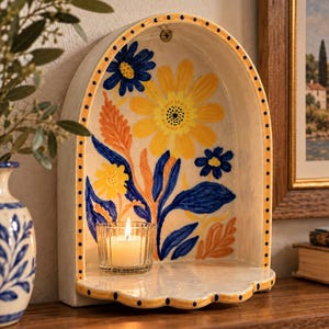May include: A cream-colored ceramic wall niche with an arched top, featuring hand-painted yellow, blue, and orange floral motifs. The edges are decorated with a yellow and blue dotted pattern. A small glass votive candle holder sits inside.
