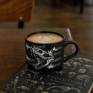 May include: A matte black ceramic mug featuring a white line-art illustration of a dinosaur skull surrounded by stars, filled with a latte topped with leaf-patterned foam, resting on a black book titled Evolution: The Story of Life on Earth.