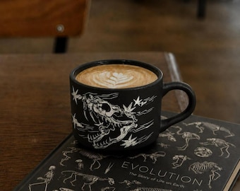 May include: A matte black ceramic mug featuring a white line-art illustration of a dinosaur skull surrounded by stars, filled with a latte topped with leaf-patterned foam, resting on a black book titled Evolution: The Story of Life on Earth.