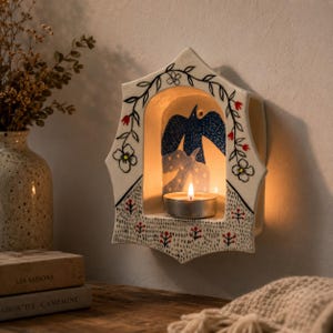 May include: A white ceramic wall-mounted candle holder with a pointed arch shape. The surface features hand-painted black line art of vines and flowers with red accents. Inside the arch, a blue bird illustration sits above a lit tea light candle.