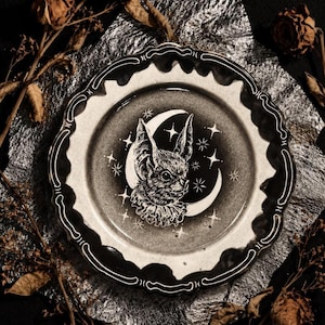 May include: A round ceramic plate with a scalloped edge featuring a black and white illustration of a bat wearing a ruffled collar centered in front of a crescent moon and stars. The plate has a dark gradient background and white line details.