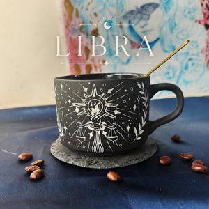 May include: A matte black ceramic mug featuring a white hand-painted design of the Libra zodiac symbol, a sunburst, and leafy branches. The mug includes a gold-toned spoon and is placed on a circular slate coaster surrounded by scattered coffee beans.