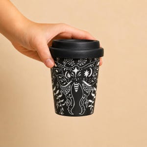 Könnte beinhalten: Ein schwarzer Reisebecher mit einem weißen Grafikdruck eines Nachtfalters, umgeben von Sternen, Monden und Blattreben. Der Becher verfügt über einen soliden schwarzen Deckel und eine konisch zulaufende zylindrische Form.