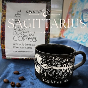 May include: A black ceramic mug featuring white etched illustrations of a bow and arrow with the word Sagittarius, positioned next to a white bag of Bush Brew Coffee with the text Ground, A Proudly Zambian Conscious Coffee, and four coffee beans.