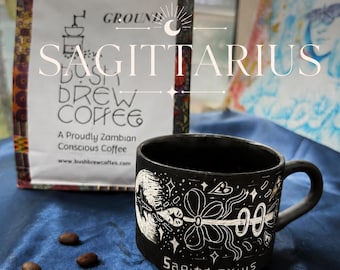 May include: A black ceramic mug featuring white etched illustrations of a bow and arrow with the word Sagittarius, positioned next to a white bag of Bush Brew Coffee with the text Ground, A Proudly Zambian Conscious Coffee, and four coffee beans.