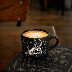 Pode incluir: Uma caneca de cerâmica preta mate com uma ilustração de linhas brancas de um crânio de animal com chifres rodeado por pequenas estrelas e linhas onduladas. A caneca está cheia de um latte com um desenho de espuma em forma de coração.