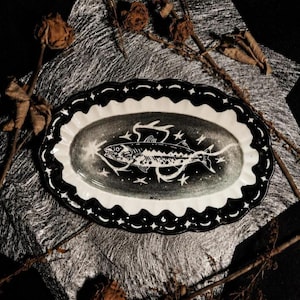 Könnte beinhalten: Ein ovales Keramiktablett mit gewelltem Rand in Schwarz und Weiß. In der Mitte befindet sich eine detaillierte Illustration eines Fisches, umgeben von kleinen Sternmotiven, während der Rand ein wiederkehrendes Muster aus schwarzen Bögen aufweist.