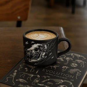 Könnte beinhalten: Ein mattschwarzer Keramikbecher mit einer weißen Strichzeichnung eines Dinosaurierschädels, Sternen und Blitzen, gefüllt mit einem Latte mit Milchschaum. Der Becher steht auf einem schwarzen Buch mit dem Titel Evolution: The Story of Life on Earth.