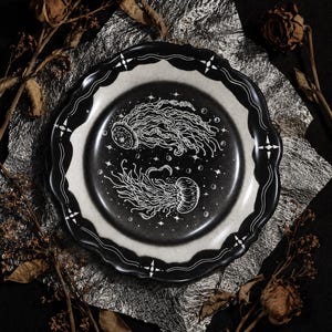 May include: A round ceramic plate with a scalloped edge, featuring a black centre decorated with two white jellyfish illustrations, small bubbles, and stars. The rim is black with white line accents and four-pointed star motifs.