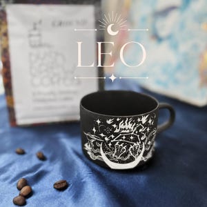 May include: A matte black ceramic mug featuring a white line art illustration of a lion, flames, stars, and a crescent moon. The word LEO is printed in large white letters above the mug, accompanied by a crescent moon and star graphic icon.