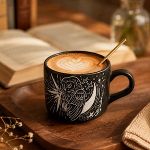 May include: A matte black ceramic mug featuring a white etched illustration of an owl perched on a crescent moon surrounded by stars. The mug is filled with a latte topped with heart-shaped foam art and contains a thin gold-toned metal spoon.