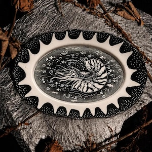 Könnte beinhalten: Eine ovale Keramikplatte mit einer schwarz-weißen Illustration eines Nautilus-Gehäuses, umgeben von kleinen Fischen und Blasen. Der Rand hat eine gewellte Kante mit einem schwarz-weißen Punktmuster, das sich vom zentralen Design abhebt.