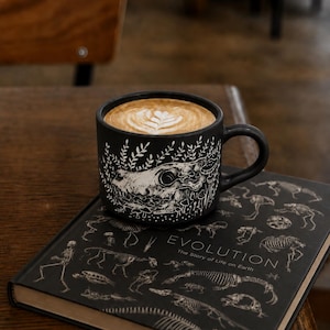May include: A matte black ceramic mug filled with latte art, featuring a white line-art illustration of an animal skull surrounded by mushrooms and foliage, resting on a black book titled Evolution: The Story of Life on Earth with skeleton graphics.