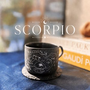 Può includere: Una tazza in ceramica nera opaca con un'illustrazione bianca disegnata a mano di uno scorpione, stelle e il simbolo zodiacale dello Scorpione. La tazza poggia su un sottobicchiere circolare in pietra scura testurizzata. Sopra c'è scritto SCORPIO.