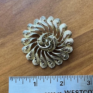 Könnte beinhalten: Goldfarbene Filigranbrosche mit Spiralmuster. Die Brosche hat einen Durchmesser von etwa 5 cm und weist aufwendige, durchbrochene Details auf. Die Nadel liegt auf einer Holzoberfläche.