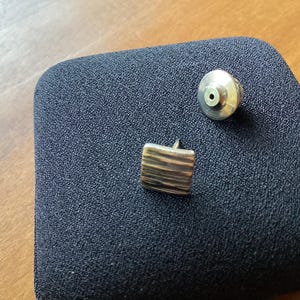 May include: A pair of square stud earrings with a textured, striped design. The earrings are made of a light-colored metal and are displayed with a matching earring back on a dark blue surface. Jewelry.