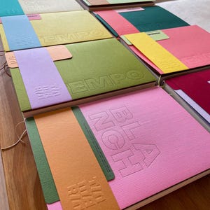 Könnte beinhalten: Eine Sammlung farbenfroher Notizbücher mit strukturierten Einbänden. Jedes Notizbuch hat eine andere Farbe, darunter Rosa, Grün, Gelb und Blau, mit geprägtem Text und farblich passenden Papierbändern. Das Wort "TEMPO" ist auf einigen Einbänden geprägt.