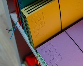 May include: A shelf displays a stack of colorful notebooks. The notebooks are in shades of yellow, purple, green, and red. The word "PLA" is visible on the yellow and purple notebooks. Each notebook has a black elastic band.