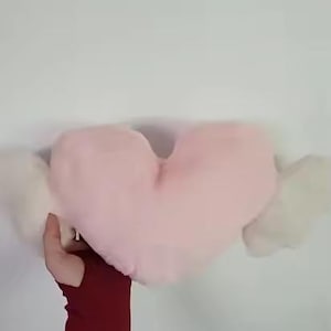 May include: A soft, plush heart-shaped pillow in a light pastel pink color, featuring two white, fluffy, wing-like extensions on either side. The pillow has a smooth, rounded texture and is held up against a plain, neutral-colored background.