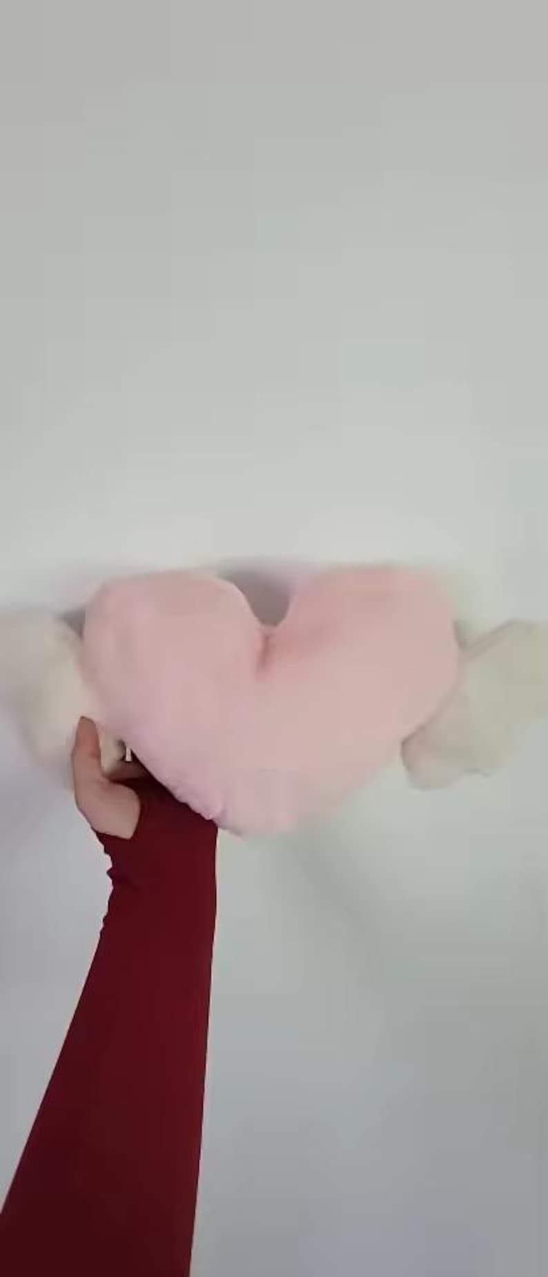 May include: A soft, plush heart-shaped pillow in a light pastel pink color, featuring two white, fluffy, wing-like extensions on either side. The pillow has a smooth, rounded texture and is held up against a plain, neutral-colored background.