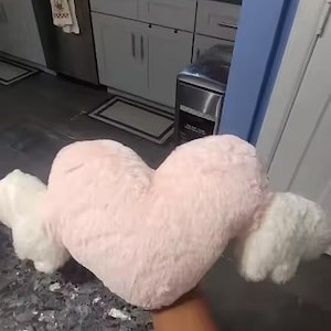 May include: A plush heart-shaped pillow in a soft light pink color featuring two white fluffy wings attached to the sides. The pillow has a textured, fuzzy surface and is held up against a kitchen background.