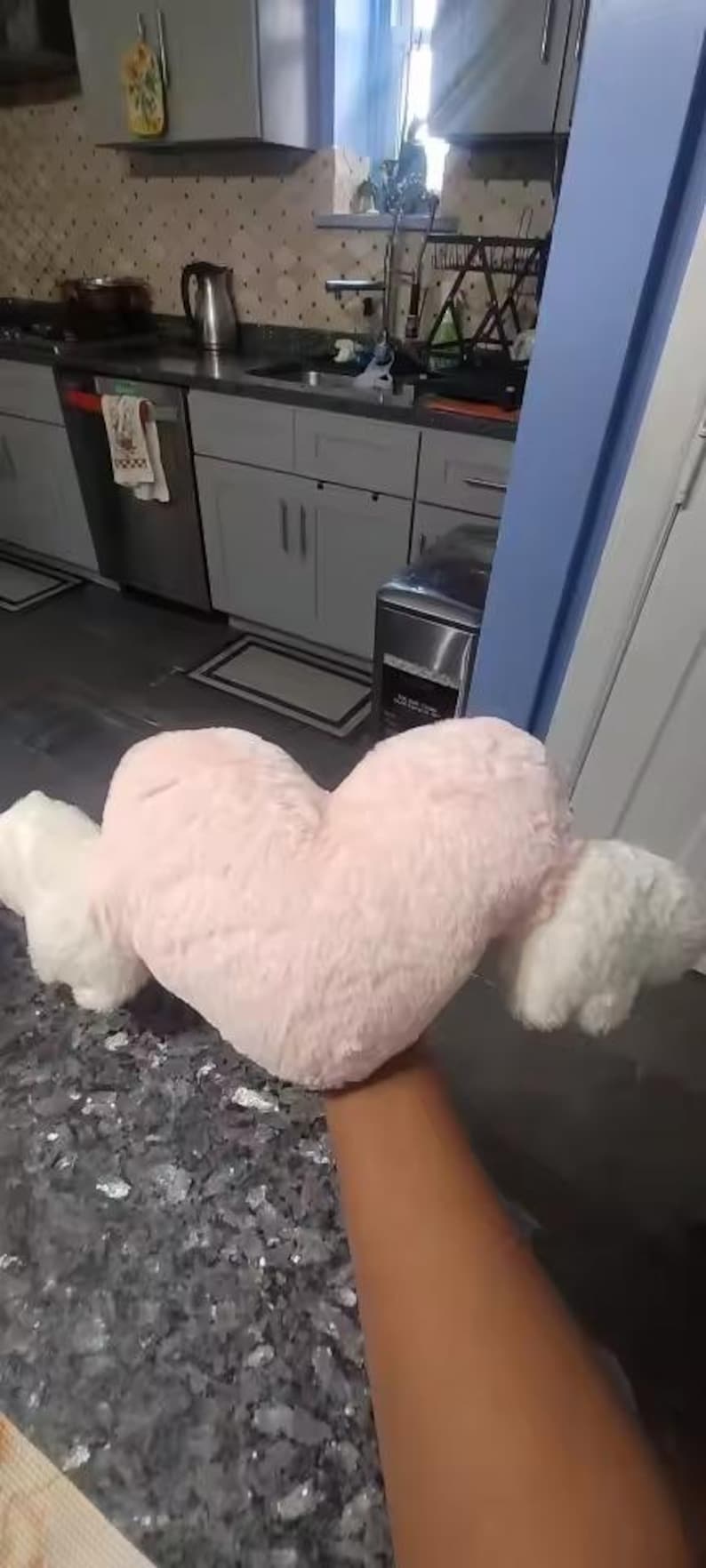 May include: A plush heart-shaped pillow in a soft light pink color featuring two white fluffy wings attached to the sides. The pillow has a textured, fuzzy surface and is held up against a kitchen background.