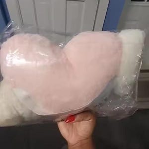 May include: A heart-shaped plush pillow with a soft, fuzzy texture, featuring a light pink center and white sides. The pillow is enclosed in a clear plastic protective bag, showcasing its rounded, stuffed form and plush material construction.