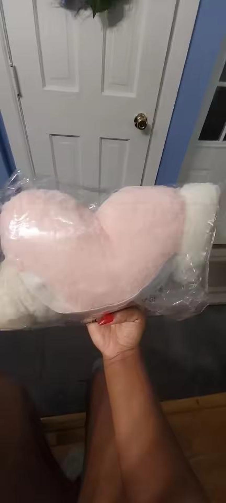 May include: A heart-shaped plush pillow with a soft, fuzzy texture, featuring a light pink center and white sides. The pillow is enclosed in a clear plastic protective bag, showcasing its rounded, stuffed form and plush material construction.