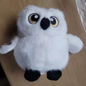 May include: A plush toy shaped like a snowy owl with soft white fur, large round eyes featuring black pupils and gold irises, a small black beak, and two black feet. The owl has a rounded body and outstretched wings, capturing a detailed and fluffy appearance.