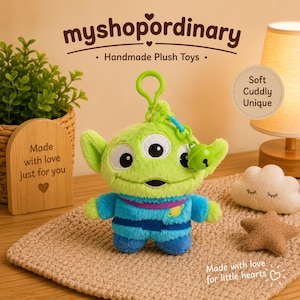 May include: A green plush alien toy with three eyes, wearing a blue and purple outfit. It has a green keychain and a small bell. The text "myshopordinary Handmade Plush Toys" is at the top. The words "Soft Cuddly Unique" are also visible.