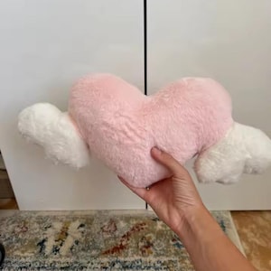 May include: A plush heart-shaped pillow in a soft pastel pink color, featuring two fluffy white wings extending from each side. The material has a textured, faux-fur finish, and the overall design is symmetrical with a rounded top and pointed base.