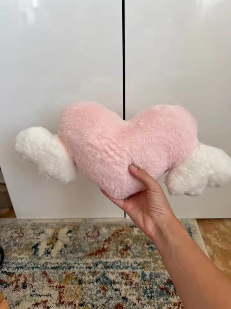 May include: A plush heart-shaped pillow in a soft pastel pink color, featuring two fluffy white wings extending from each side. The material has a textured, faux-fur finish, and the overall design is symmetrical with a rounded top and pointed base.