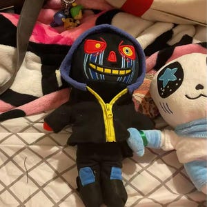 May include: A black plush doll with a blue-lined hood and yellow zip. The doll has a detailed face with red and blue accents. Black trousers with blue knee patches are visible. A panda-faced plush is also in the image.