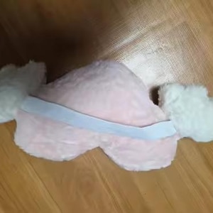 May include: A plush heart-shaped pillow in a soft light pink color featuring two white fluffy wings attached to the sides. A wide white elastic band is stretched horizontally across the center of the pink heart, connecting the two white wings.