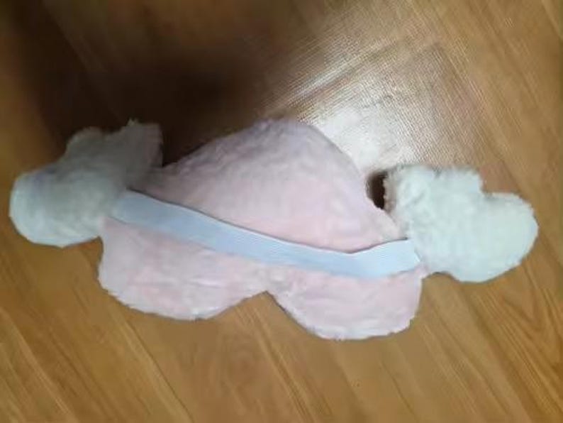 May include: A plush heart-shaped pillow in a soft light pink color featuring two white fluffy wings attached to the sides. A wide white elastic band is stretched horizontally across the center of the pink heart, connecting the two white wings.