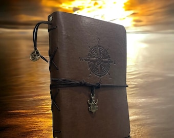 May include: A brown leather-bound journal featuring a debossed compass design on the front cover. The journal is secured with a dark leather cord wrap and includes a metal pentacle charm and a small metal mask charm attached to the binding.