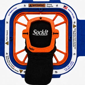 May include: A blue and orange 5.5 inch Mighty Hoop embroidery frame with a white border containing warning text and a US flag. A black sock is secured in the centre by an orange frame piece labelled Sockit. Metal plates are attached to the sides.