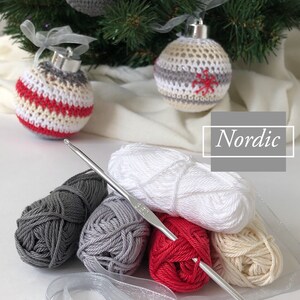May include: Handmade Christmas baubles in red, white, and grey, with silver tops and ribbons, are displayed near a Christmas tree. The image includes yarn, a crochet hook, and the word "Nordic".