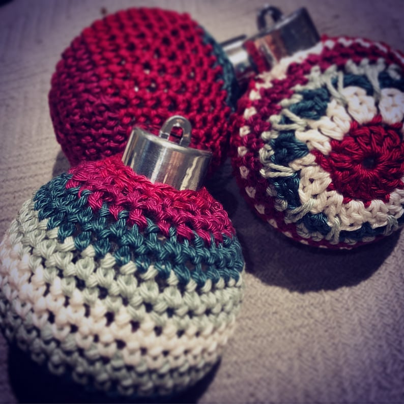 May include: Close-up of three handmade Christmas baubles. The baubles are crocheted with red, white, green, and pink yarn. Each bauble has a silver metal cap. The baubles are resting on a neutral-coloured surface.