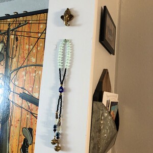 May include: A white wall with a brass elephant head wall decor, a black and white beaded necklace with blue evil eye charms and bells, and a metal mail holder with postcards inside.